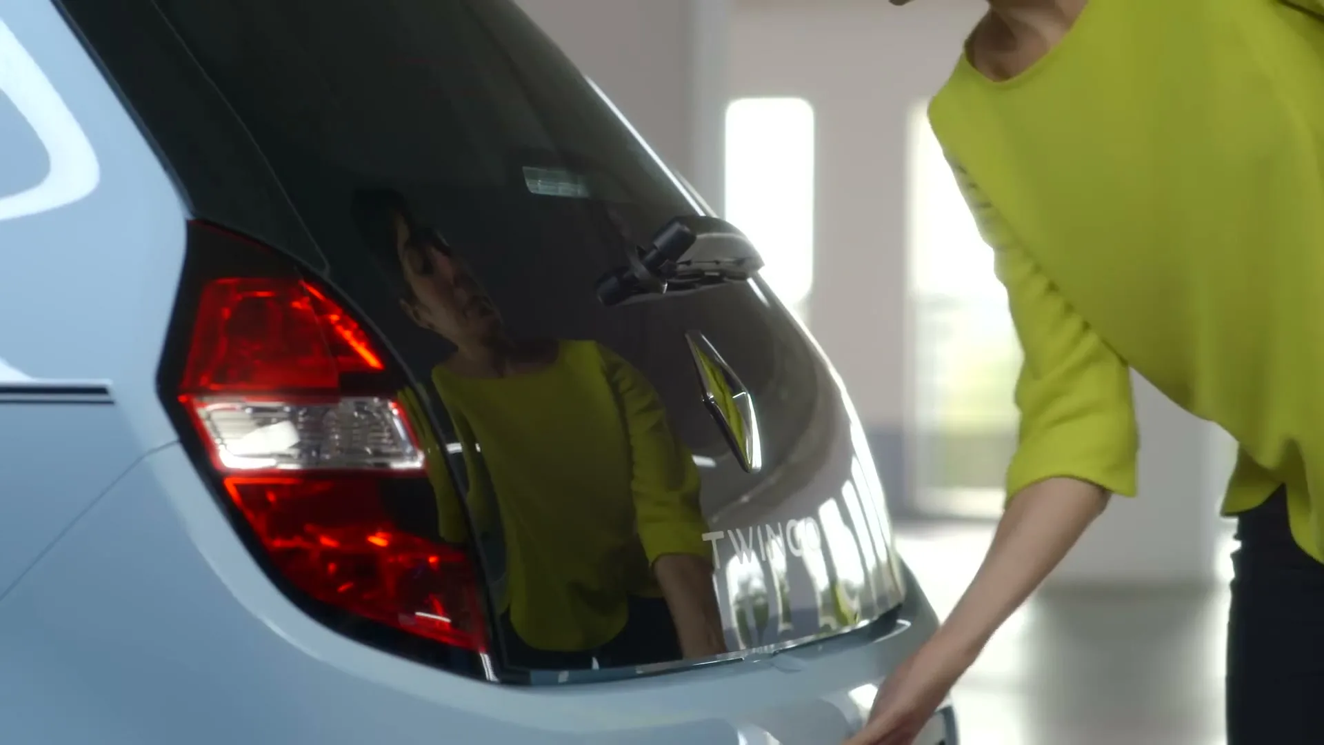 A woman in a bright yellow top opens the trunk of a light blue Renault Twingo in a brightly lit, modern garage.