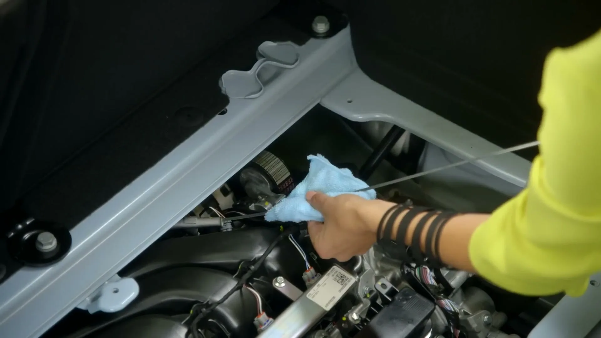 A woman's hands in a bright yellow top check the oil in a car engine, wiping the dipstick with a blue cloth.