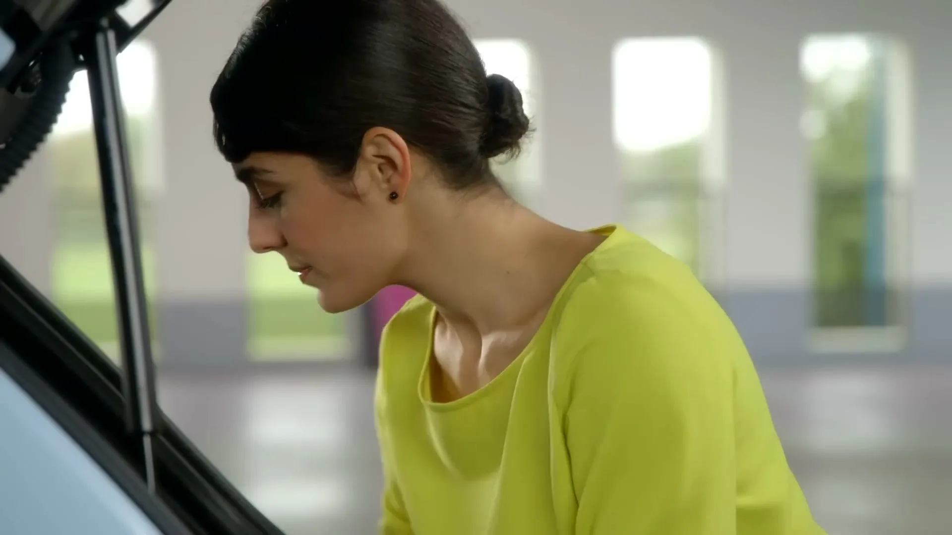 A woman with dark hair in a bun and a lime green top looks into the trunk of a light blue car in a bright, modern garage.