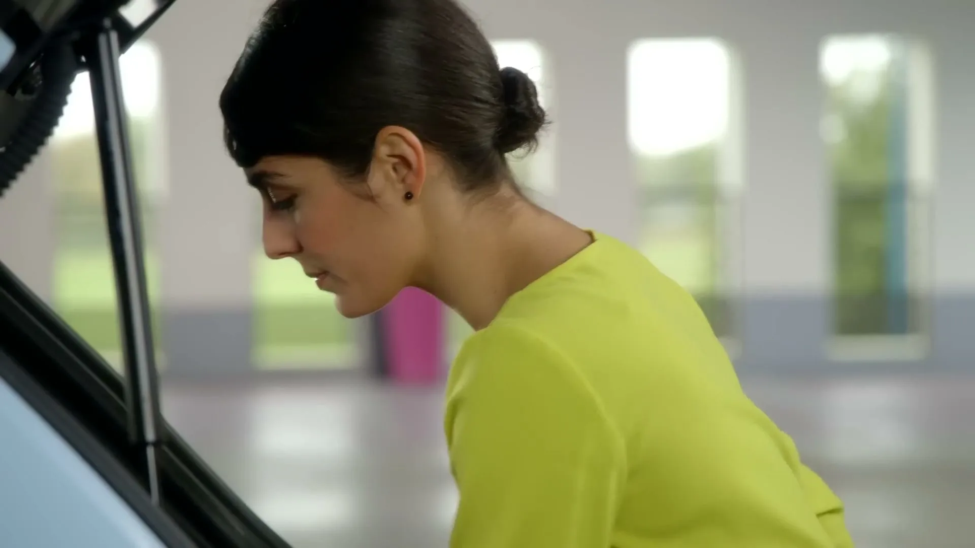 A young woman with dark hair in a bun, wearing a bright yellow top, looks down into the open trunk of a light blue car in a bright, modern garage.