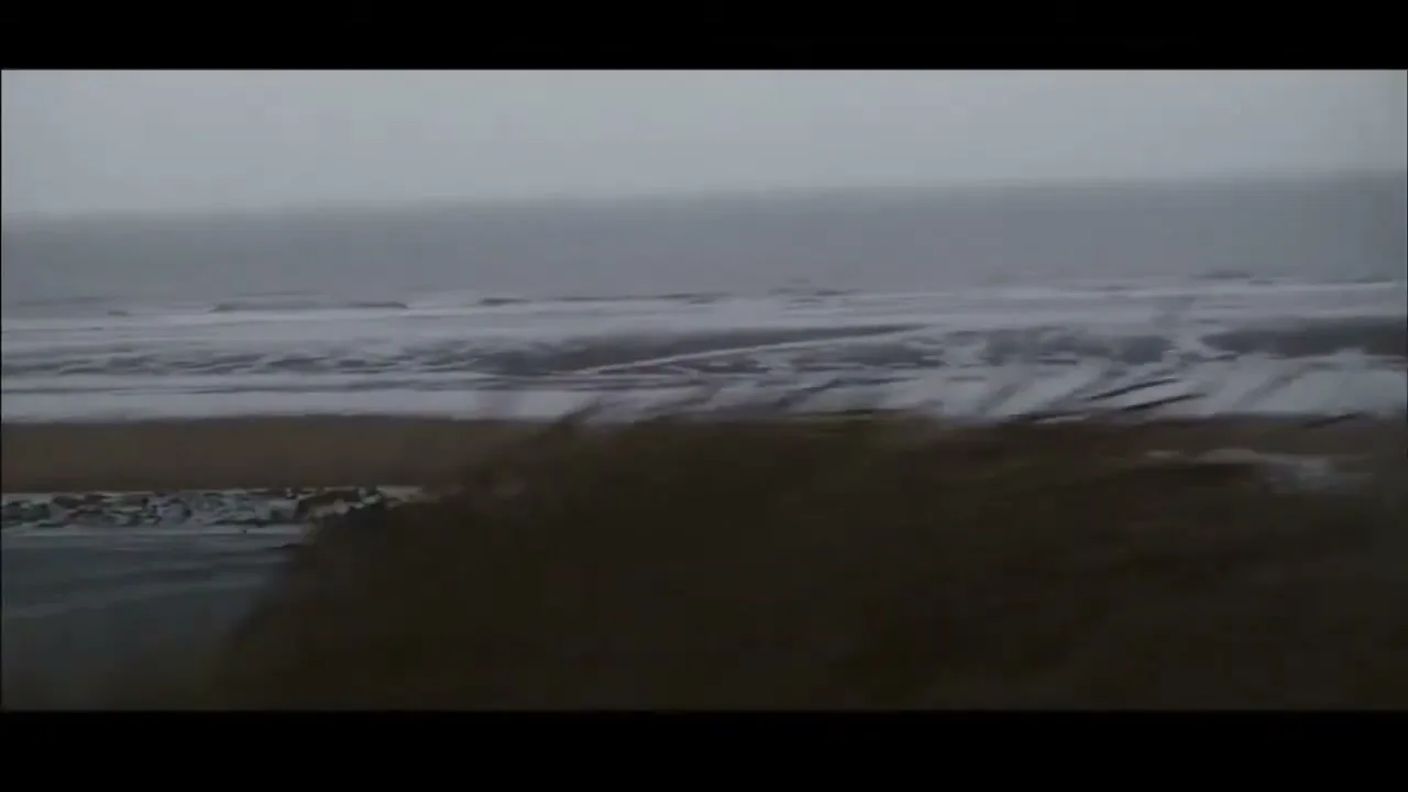 A wide shot of a desolate, snow-covered beach with dark, choppy waves under a grey, overcast sky.