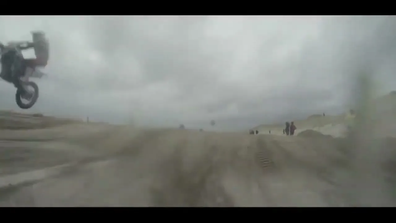 A motocross rider in red and white gear performs a wheelie on a dirt bike on a sandy beach under a cloudy sky, with spectators in the distance.