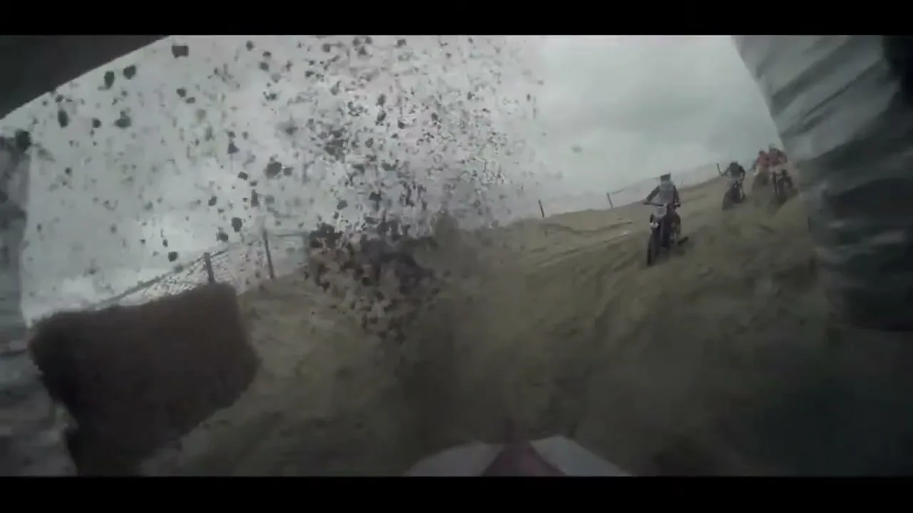 A first-person view from a motocross bike shows dirt and debris flying as other riders race on a sandy track under an overcast sky.