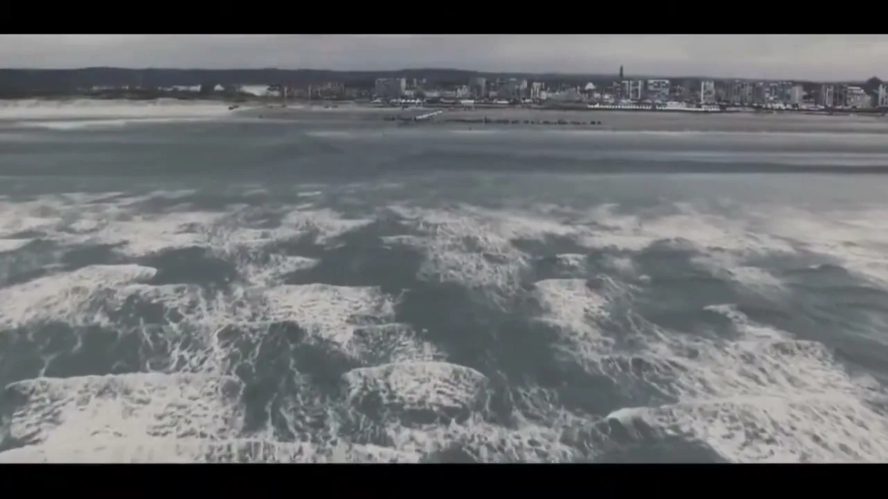 An aerial view shows a vast expanse of ocean with white waves crashing, and a distant city skyline under a cloudy sky.