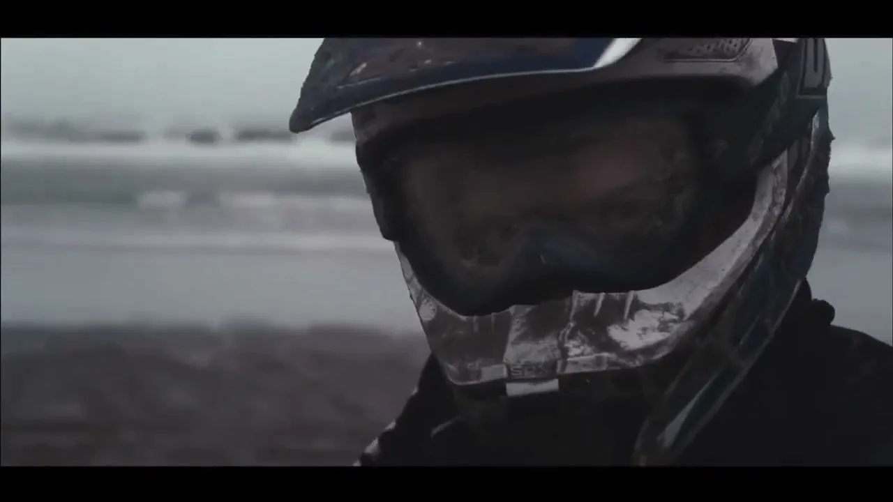 A person in a dirty motocross helmet and goggles looks down, with a blurry beach and ocean in the background under a cloudy sky.
