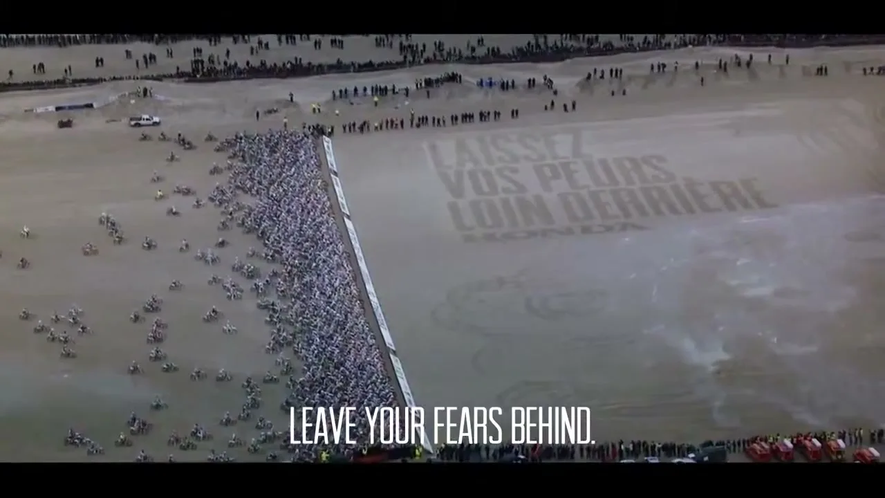 An aerial shot shows hundreds of motocross riders lined up on a sandy beach, with a large message written in the sand next to them.