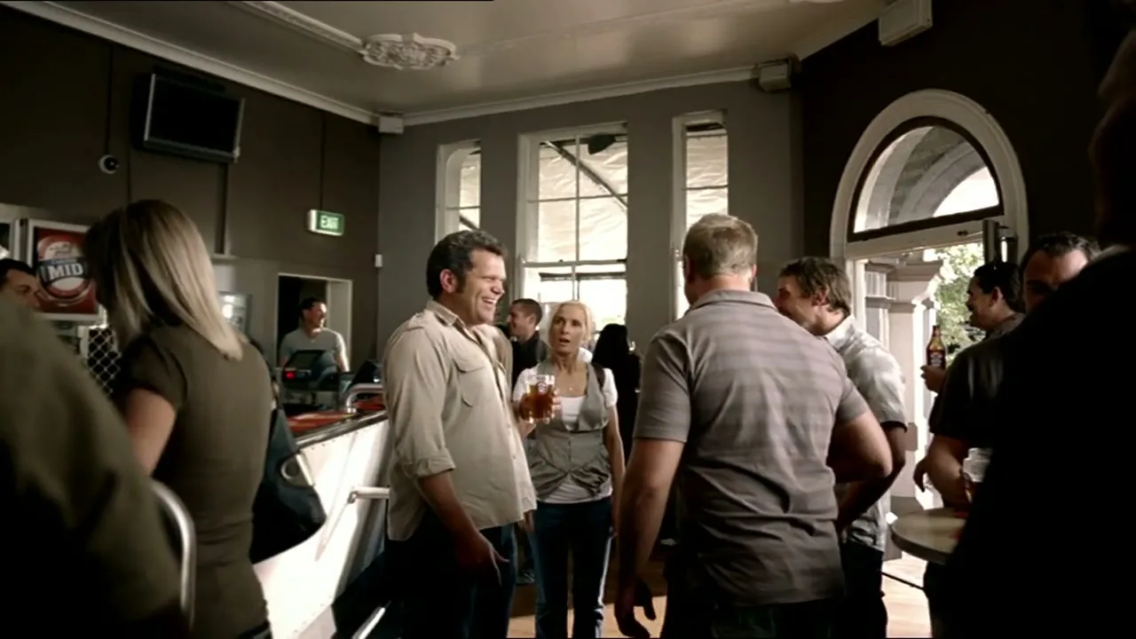 A man in a beige shirt laughs with a group of people in a busy pub, while a woman holds a drink and smiles in the background.