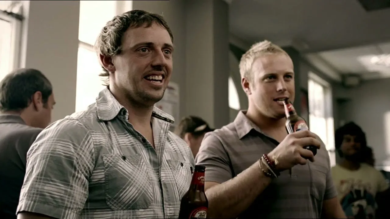 Two men in their 20s-30s, one with a plaid shirt and the other with a grey polo, are smiling and drinking beer in a bar.