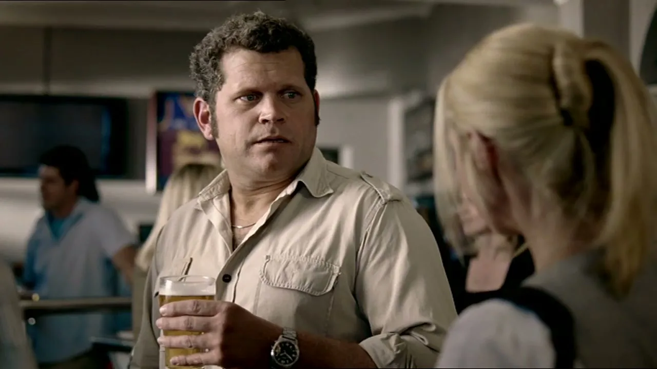 A man with curly hair and a beige shirt holds a beer and looks intently at a blonde woman in a pub.