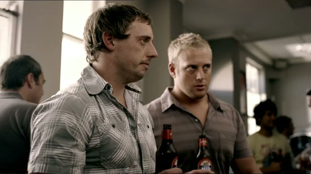 Two men, one with dark hair and a plaid shirt, the other with blonde hair and a striped shirt, stand at a bar, holding beer bottles and looking intently at something off-screen.