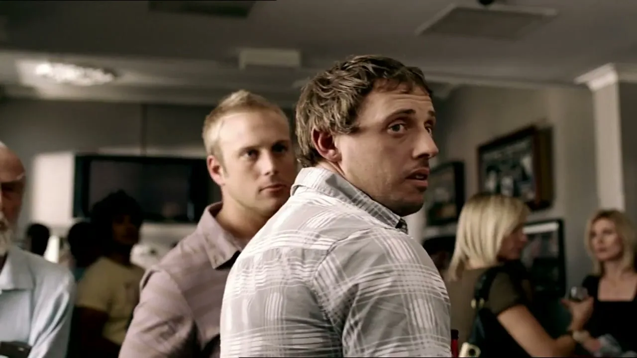 A man with curly hair in a plaid shirt looks over his shoulder, surprised, in a crowded indoor setting with other people in the background.