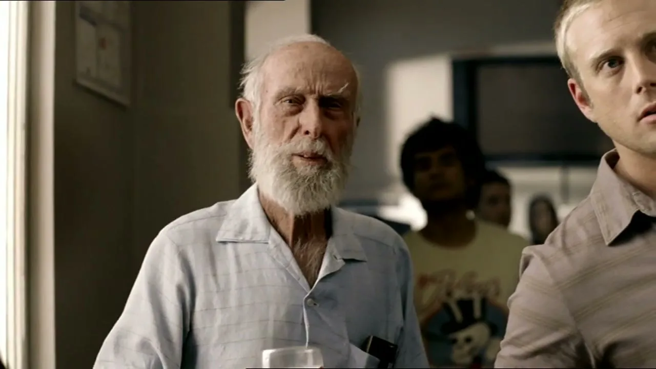An elderly man with a white beard holds a beer and looks surprised, standing in a crowded room with other people in the background.