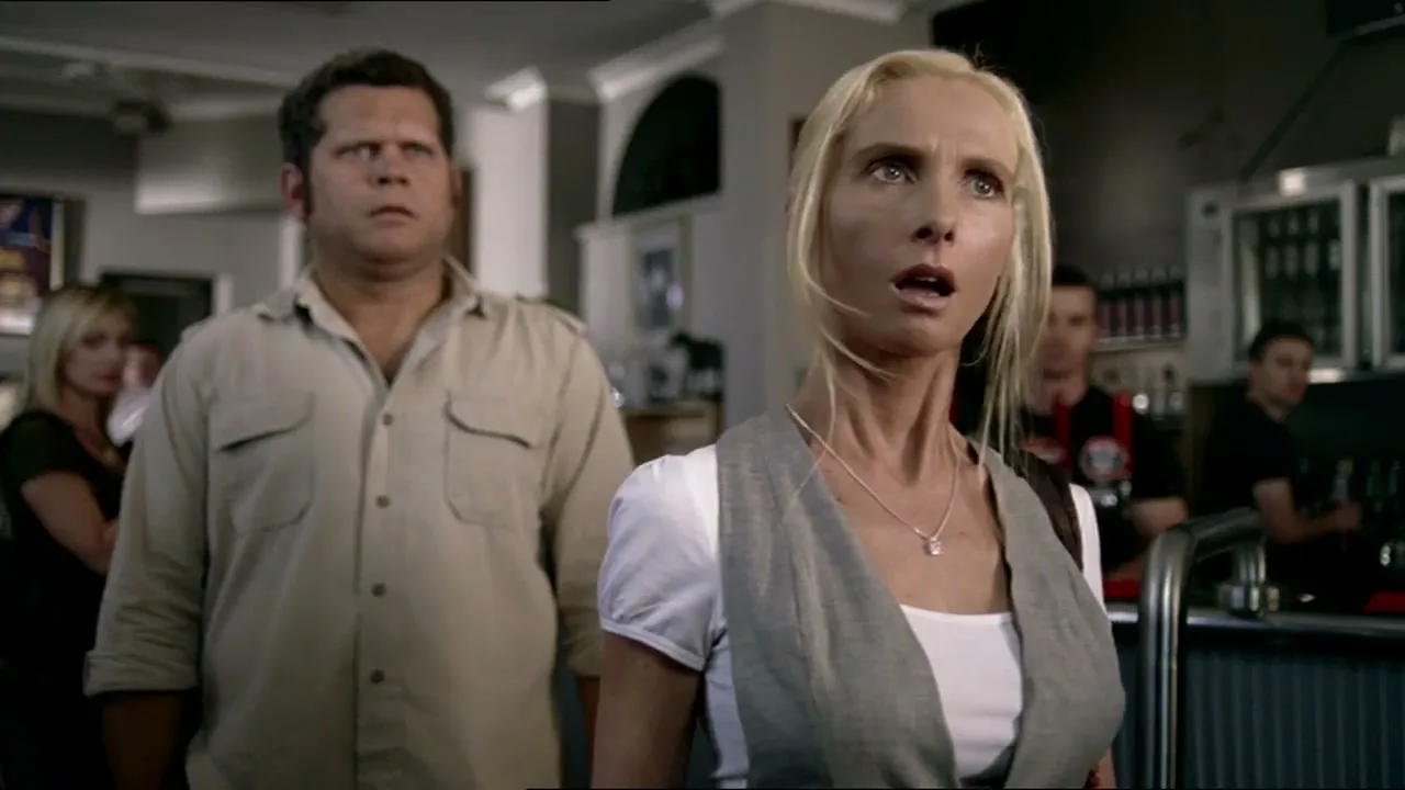 A blonde woman in a white shirt and grey vest looks shocked, standing in a busy bar with a man in a beige shirt behind her.