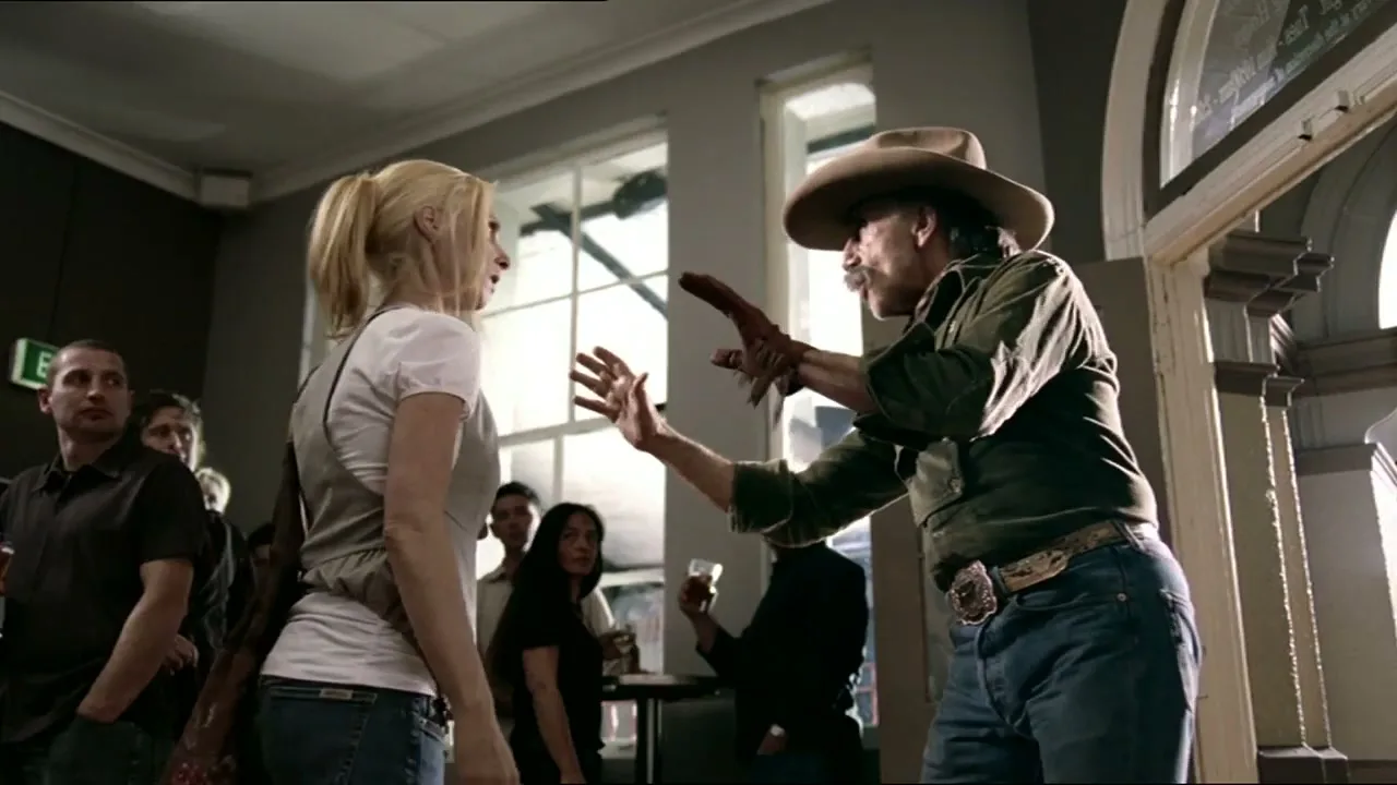 A man in a cowboy hat and a woman in a white t-shirt are talking in a crowded bar, with other patrons in the background.