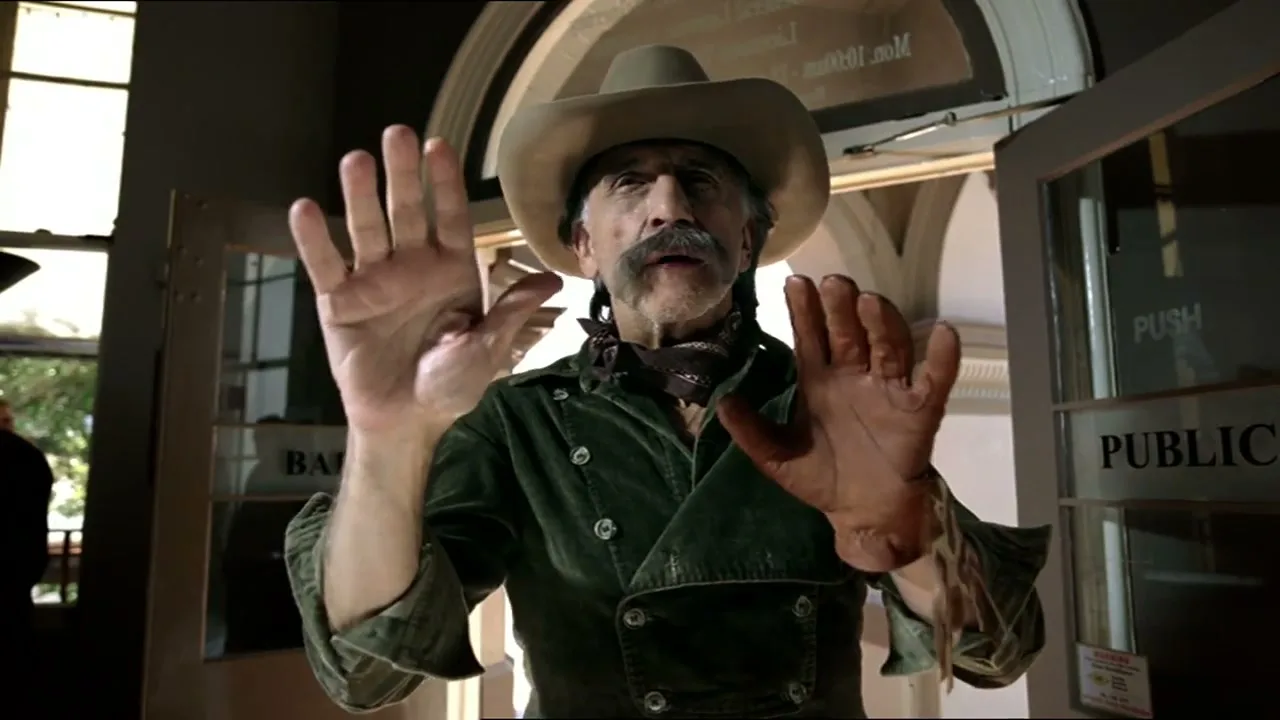 A man with a mustache and cowboy hat, wearing a green jacket, holds his hands up as if pushing something away, in a public building.