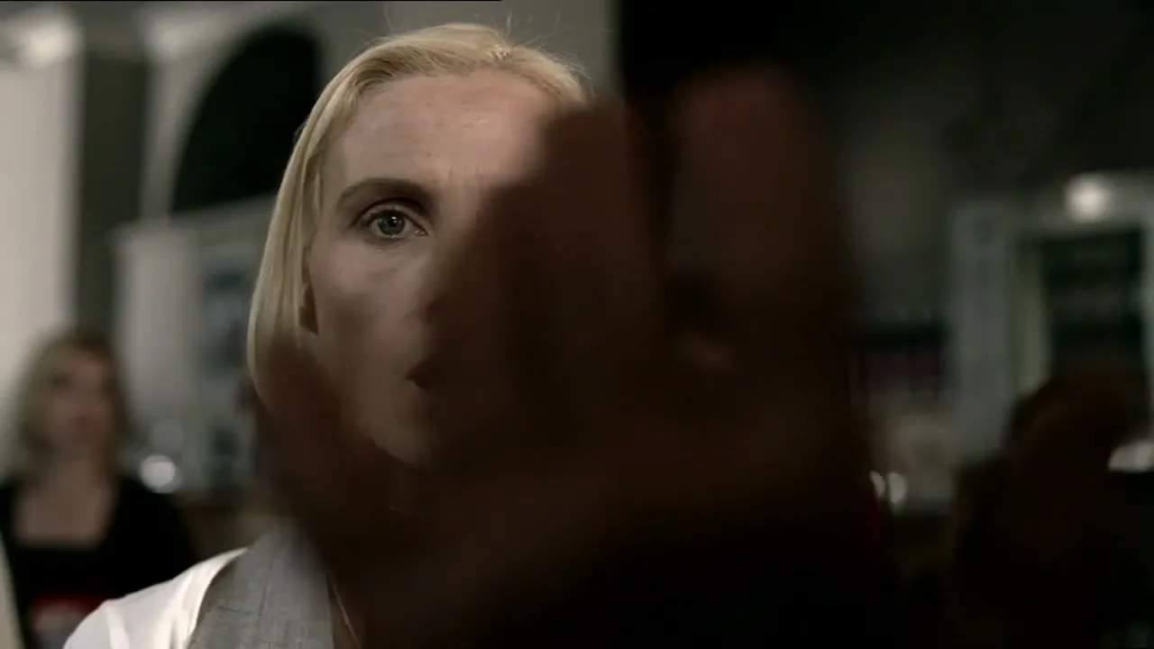 A blonde woman with wide eyes and an open mouth looks surprised as a hand moves in front of her face in an interior setting.