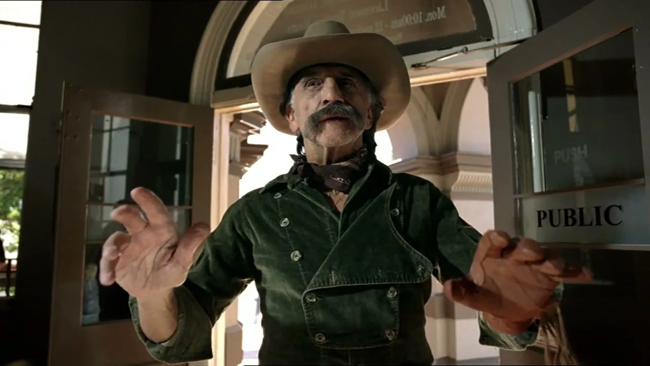 A man in a cowboy hat and green jacket stands with his hands up, looking surprised in a doorway.