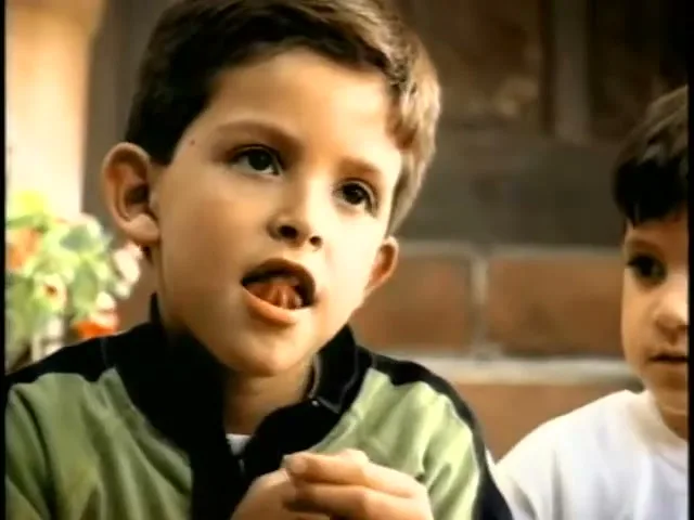 A young boy in a green and black jacket wiggles his loose tooth with his finger, while another boy watches him intently in an outdoor setting.