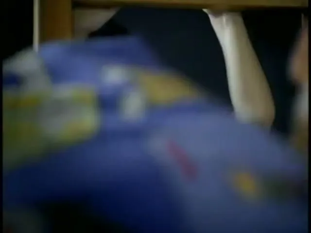 A young boy with dark hair and a grey shirt looks up from his bed, holding onto the wooden frame of a bunk bed in a dark bedroom.