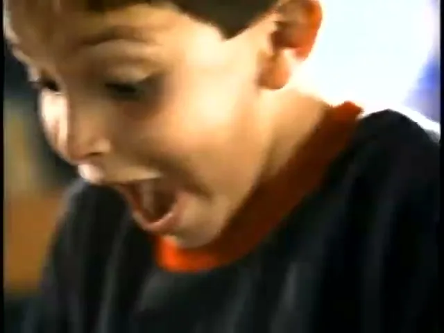 A young boy with dark hair and a blue shirt with a red collar looks down with his mouth open in surprise.