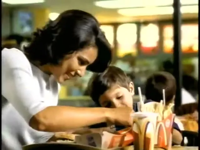 A mother and her young son are sitting at a table in a McDonald's restaurant, with the mother helping the son with his food.