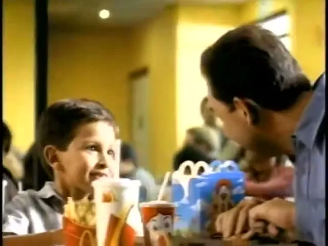 A father and son sit at a table in a McDonald's restaurant, with the boy looking at his father and smiling while eating fries.