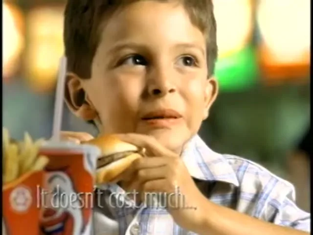A young boy in a plaid shirt eats a hamburger and fries at a McDonald's, looking up and smiling with a missing front tooth.