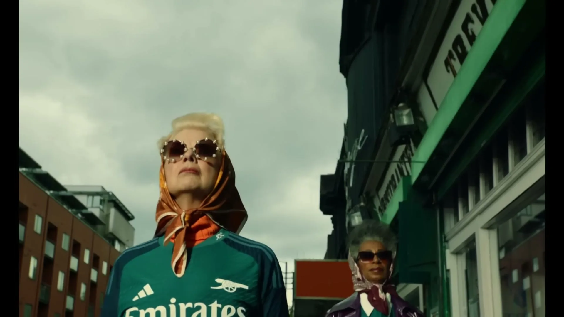 Two older women, one with blonde hair and the other with grey hair, wear headscarves, sunglasses, and Arsenal jerseys as they walk down a city street.