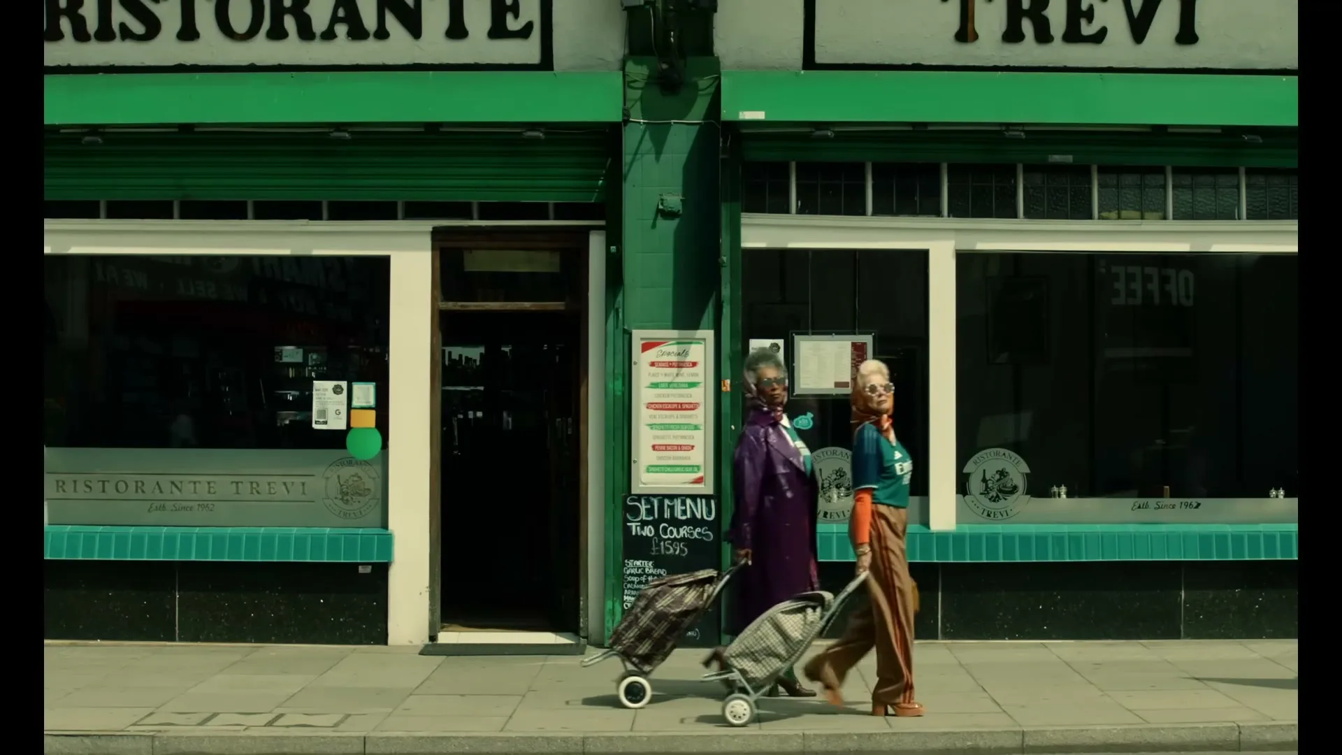 Two older women, one in a purple coat and the other in a teal shirt, walk past a green Italian restaurant with shopping carts on a sunny day.
