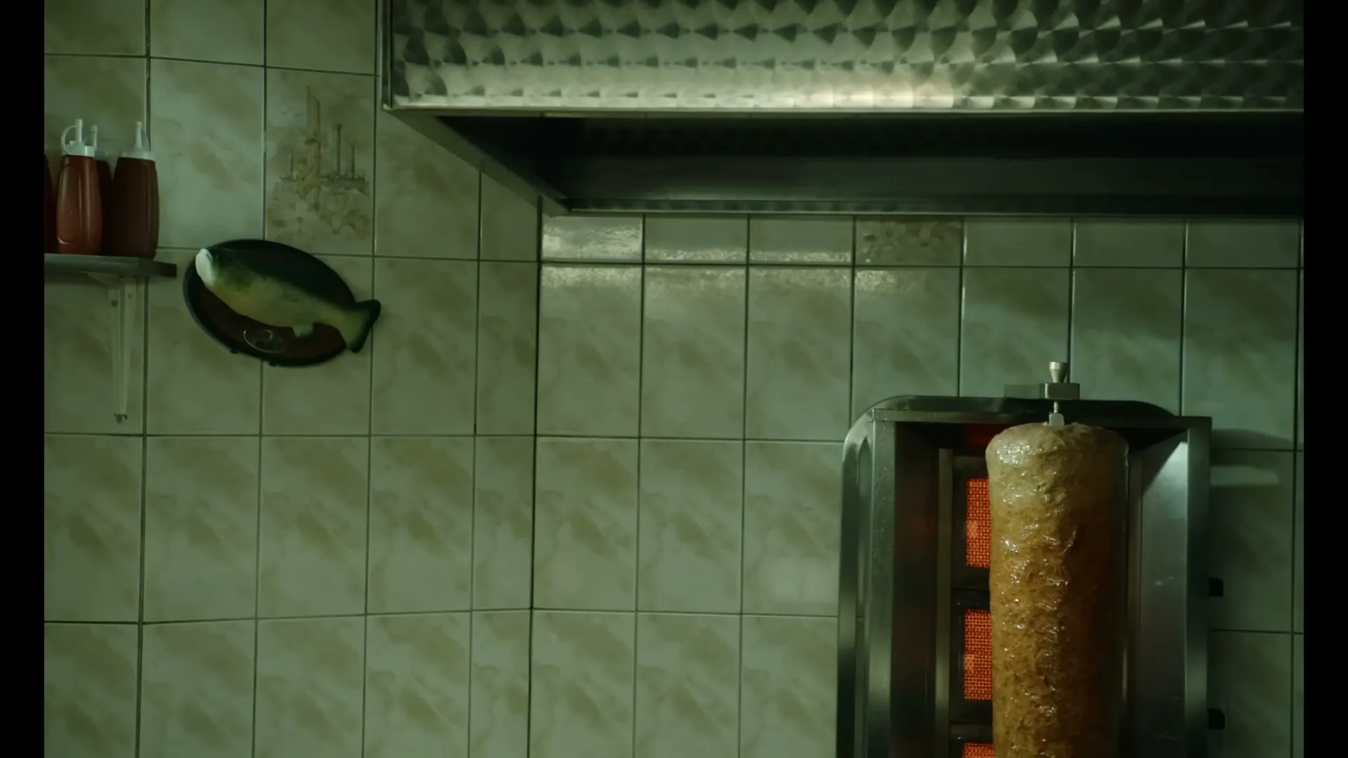 A close-up shot of a rotating kebab skewer cooking in a dimly lit, tiled kitchen, with a fish decoration on the wall.