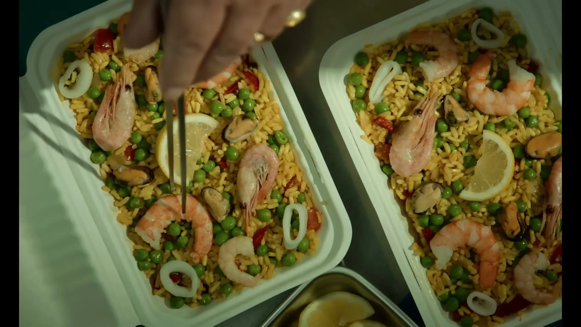 A person's hands are seen from above, placing a lemon wedge into a tray of paella with shrimp, mussels, and peas.