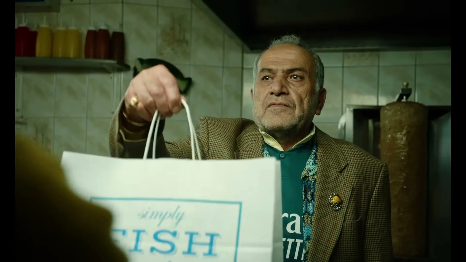 A middle-aged man with a beard, wearing a brown jacket and a teal shirt, hands over a white paper bag in a tiled kitchen.