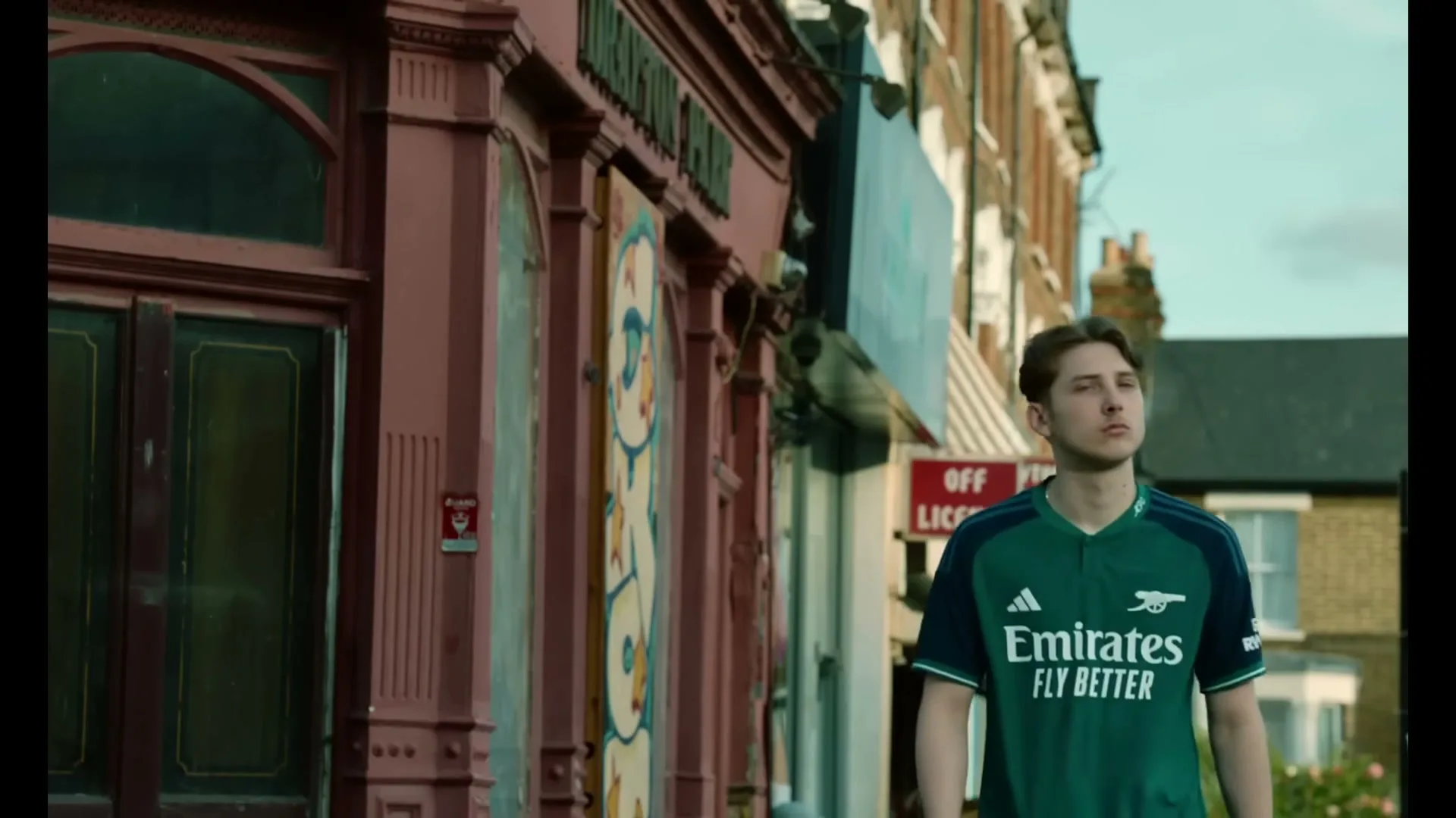 A young male in his 20s, wearing an Arsenal football jersey, walks along a street with old buildings and shops, looking up at the sky.