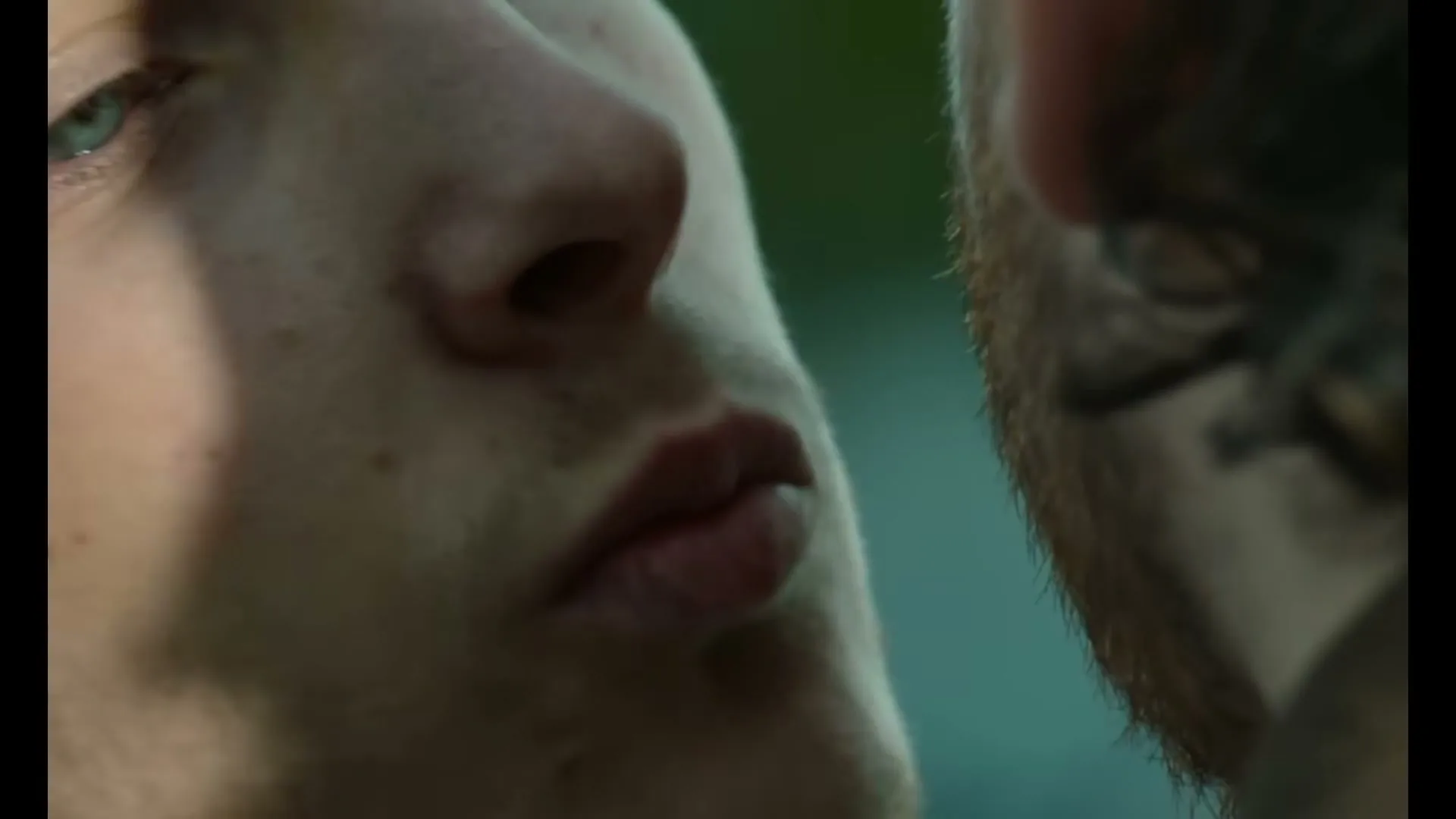 A young man with blue eyes and light stubble looks intently at another man's ear, who has a beard and dark hair, in a close-up shot.