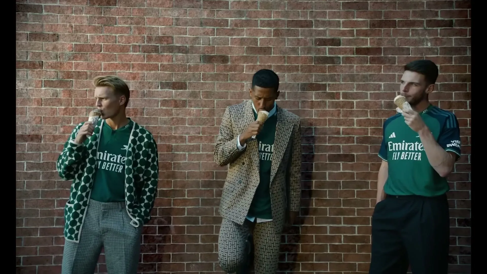 Three male soccer players, wearing green jerseys and stylish outfits, eat ice cream cones while leaning against a brick wall outdoors.
