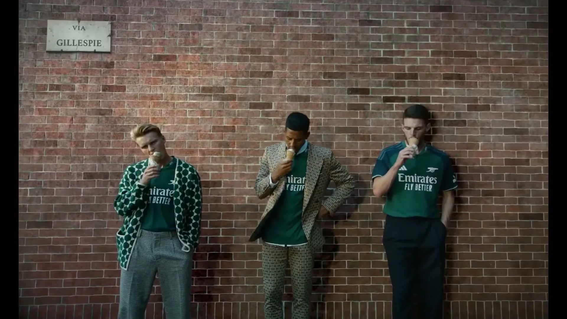Three male soccer players in stylish outfits and green jerseys stand against a brick wall, eating ice cream cones on a street.