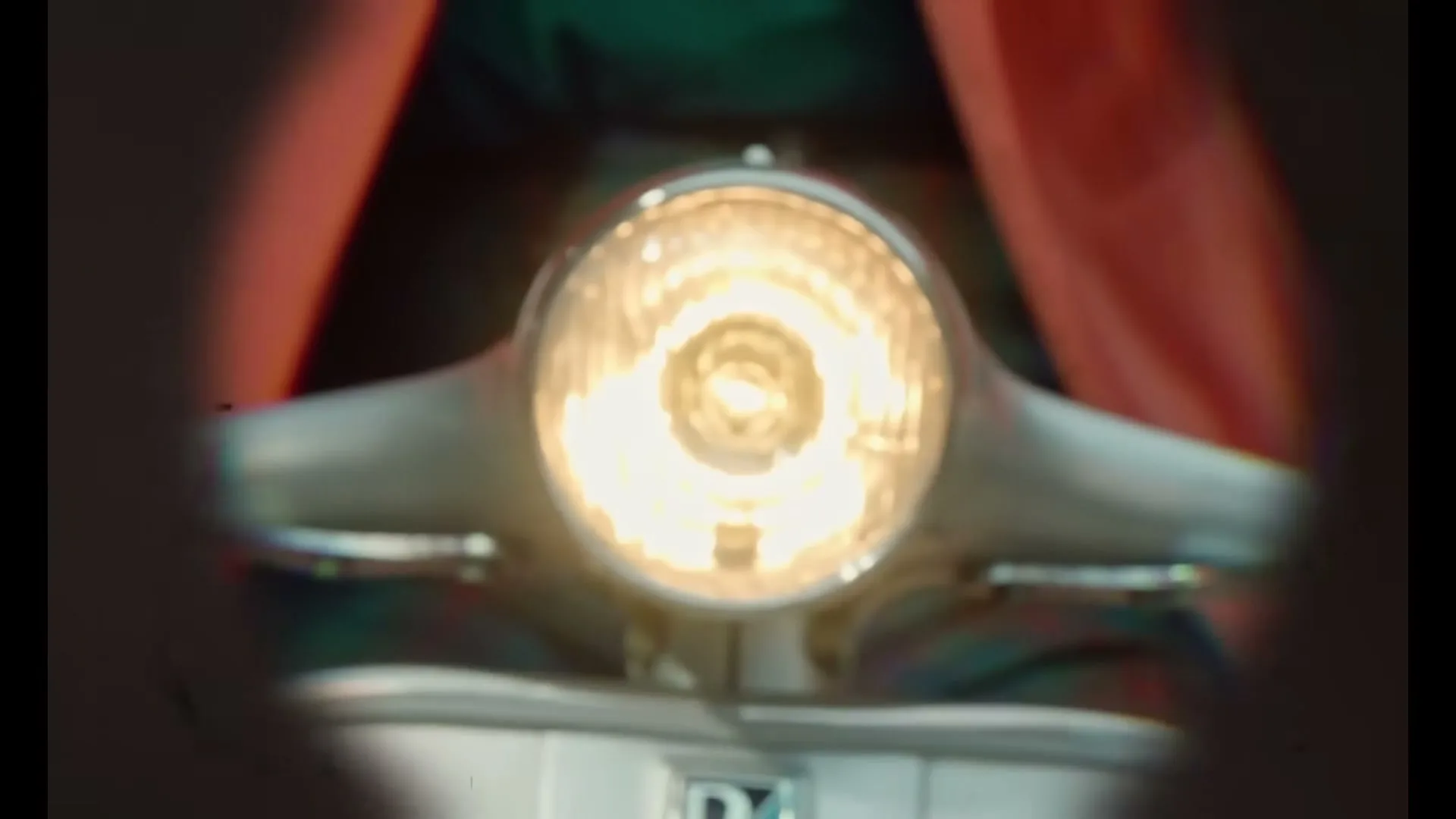 A bright, glowing headlight of a vintage scooter is seen through a dark, circular vignette, with red fabric in the background.