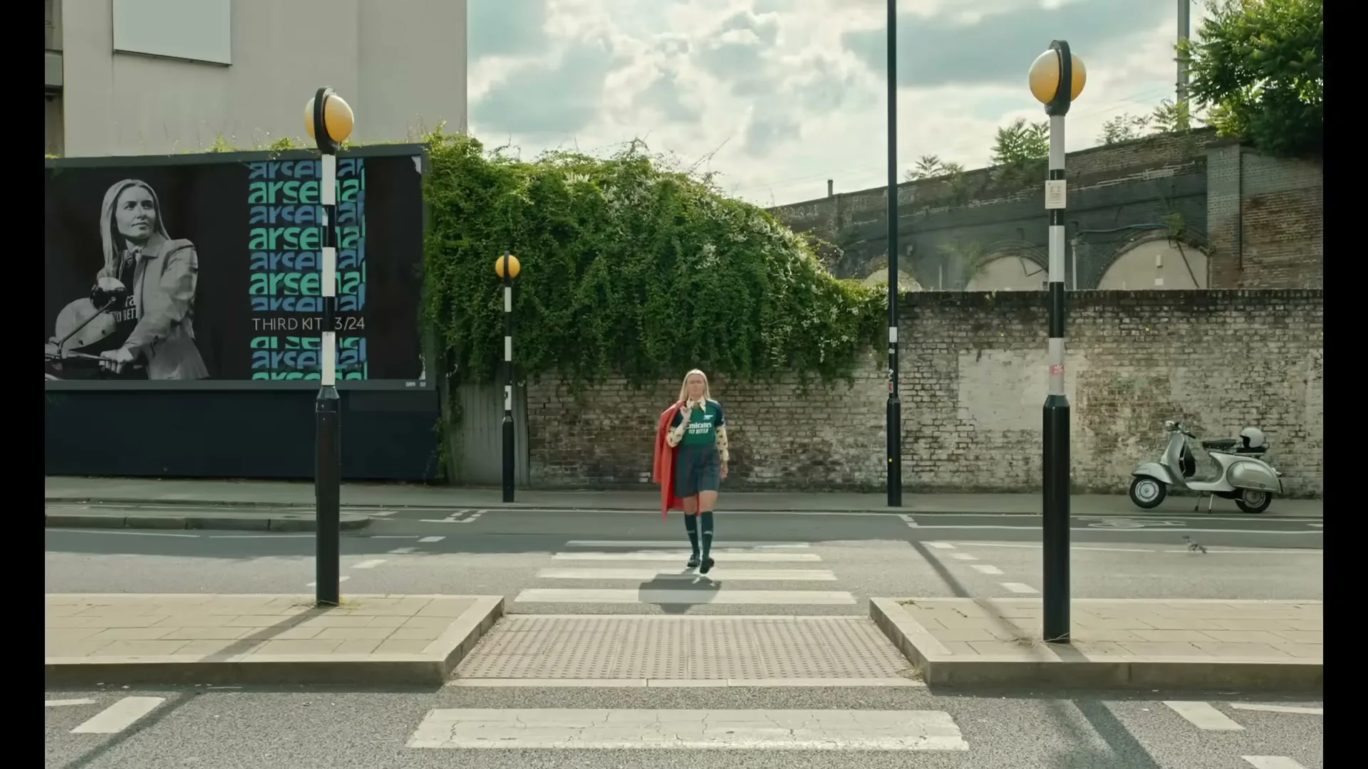 A young woman in a green jersey and red cape walks across a crosswalk in a city, with a billboard and a scooter in the background.