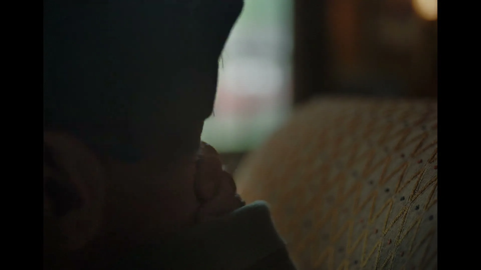 A man in his 60s or older, wearing a dark cap, is seen from behind, looking down, with a patterned cushion beside him in an interior setting.