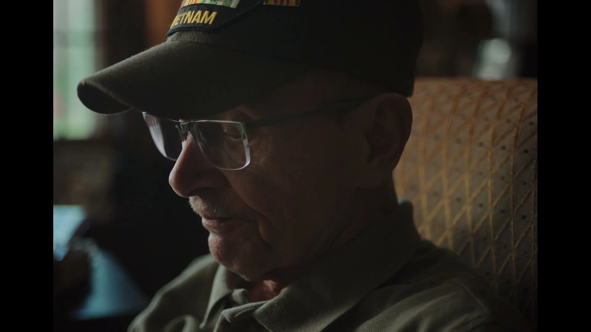 A senior man with glasses and a "Vietnam" cap sits in a chair, looking down and speaking softly in a dimly lit room.