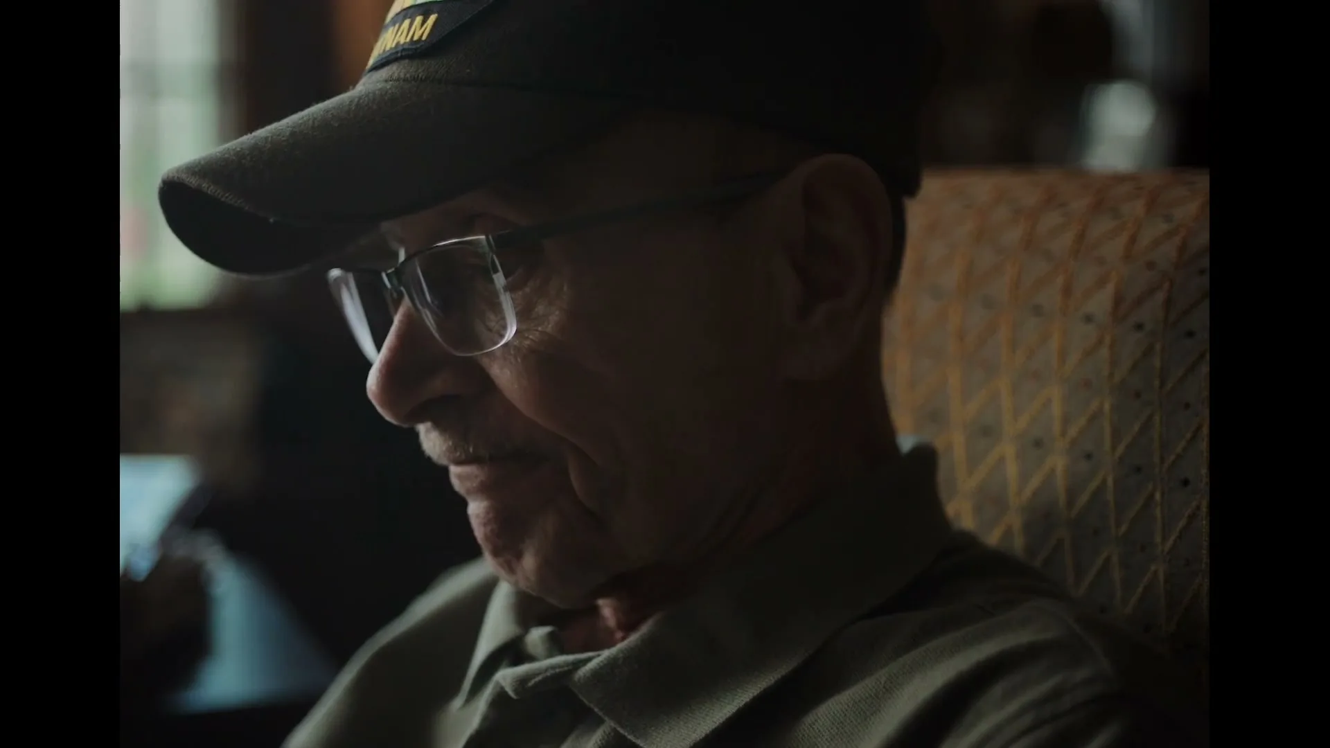 A senior man wearing a "Vietnam" cap and glasses sits in a patterned armchair, looking down and smiling slightly in a dimly lit room.