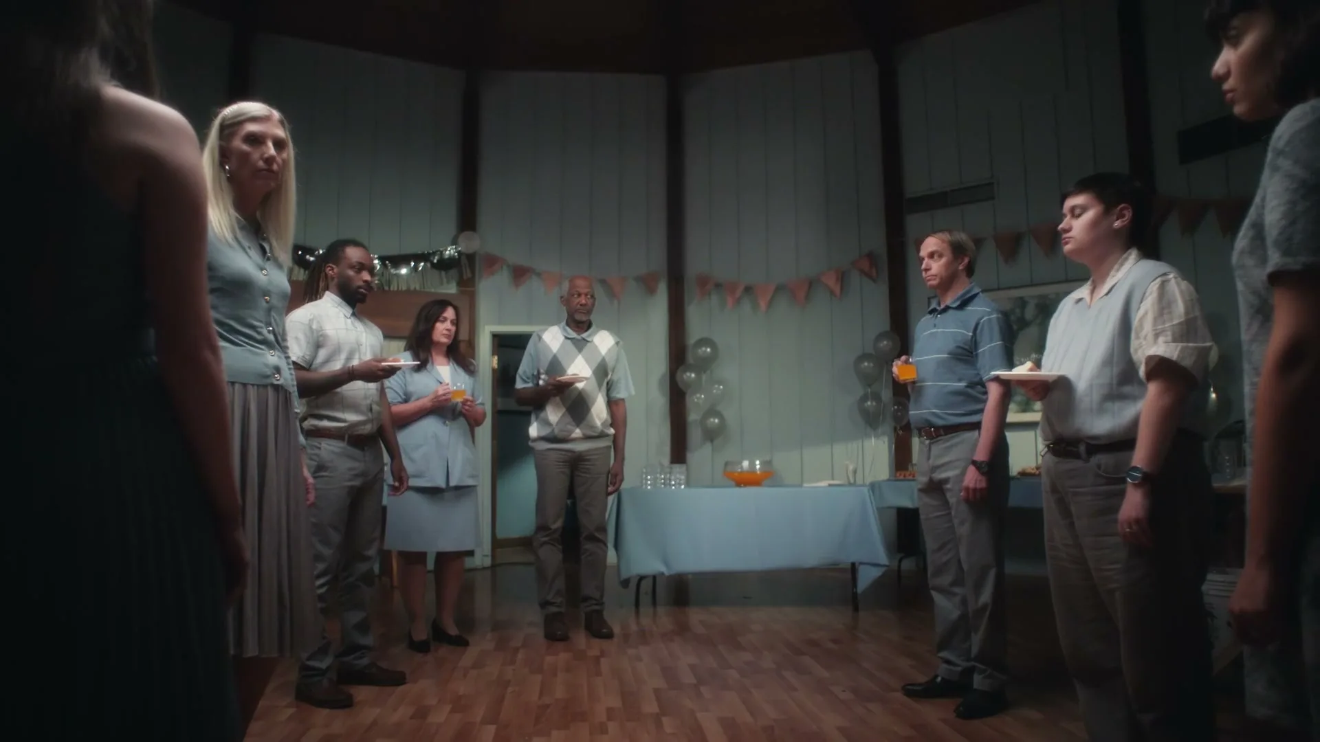A diverse group of people, dressed in muted colors, stand awkwardly in a circle at a dimly lit party, holding plates and drinks.
