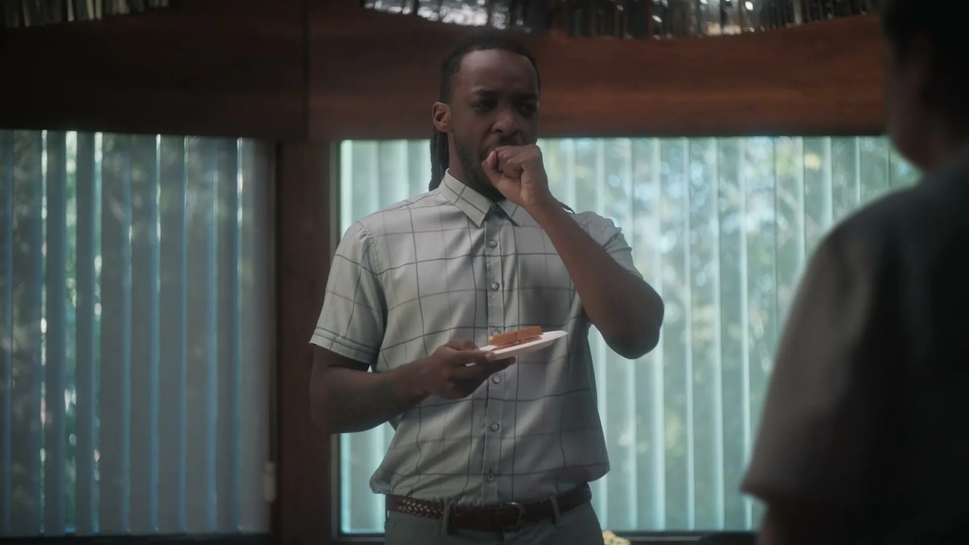A man with dreadlocks in a plaid shirt holds a plate with food, looking disgusted, then smiles at someone off-screen at an indoor party.