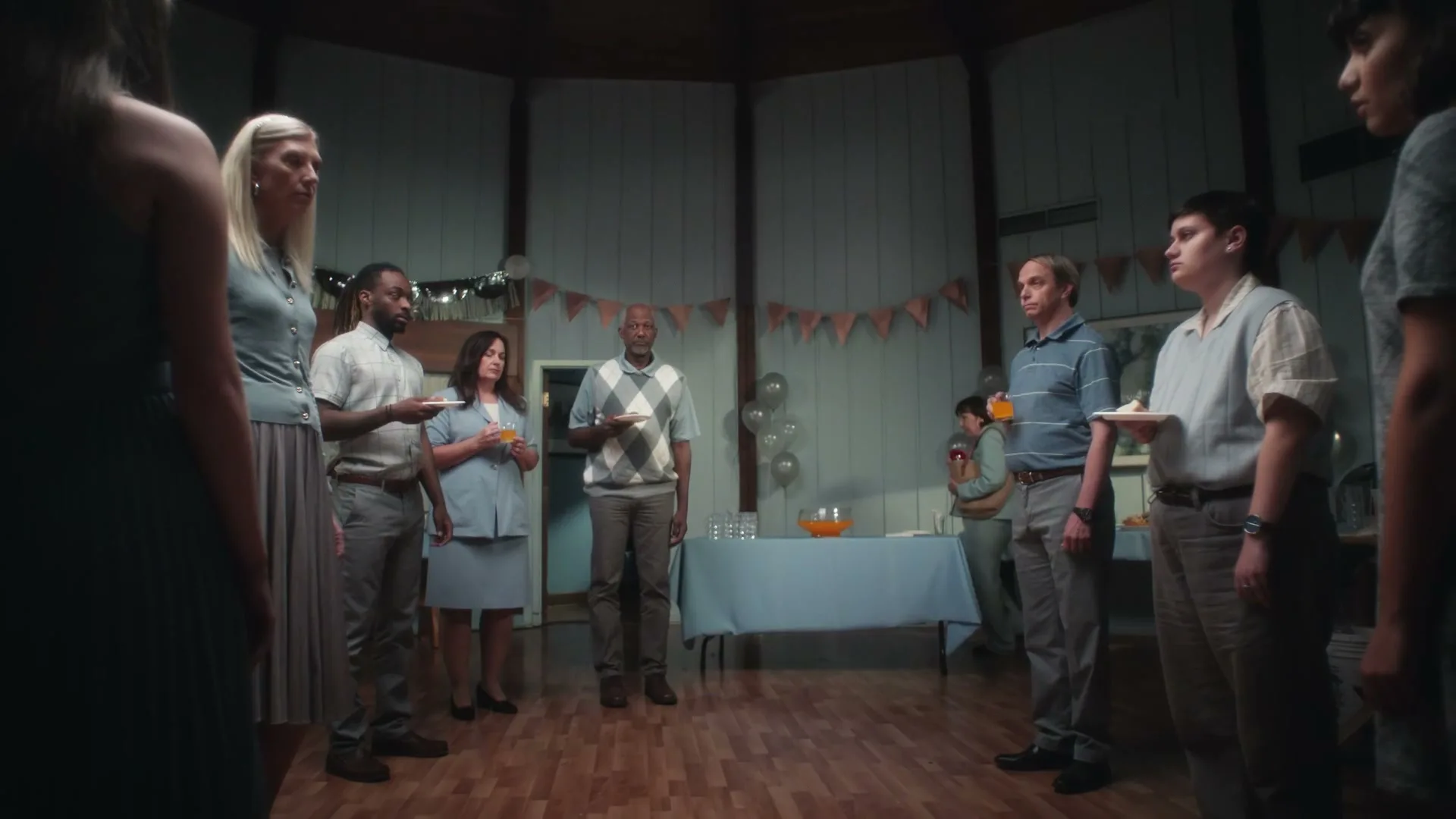 A diverse group of people, dressed in light blue and grey attire, stand in a circle in a dimly lit room, holding drinks and plates at a party.