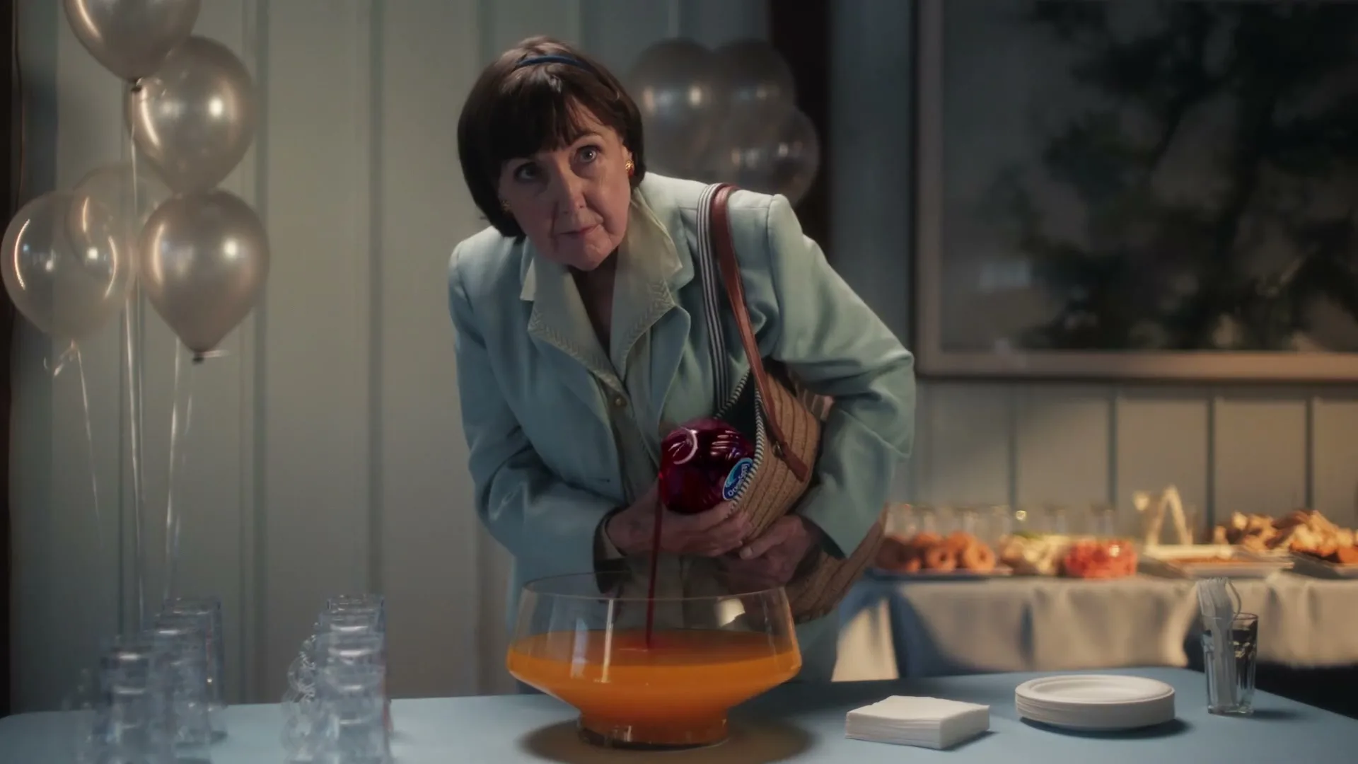 A woman in her 60s, wearing a light blue suit, discreetly pours Ocean Spray cranberry juice into a punch bowl at a party.