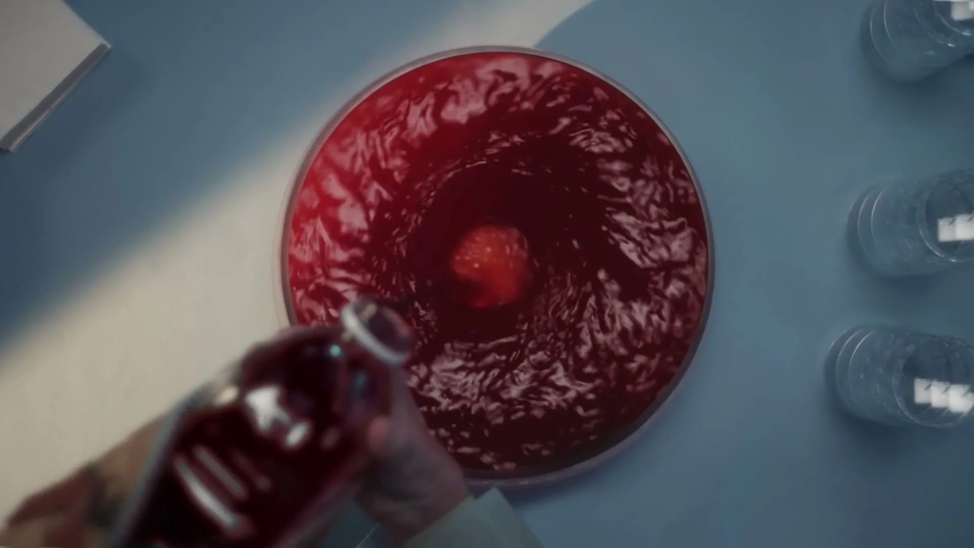 A person's hand pours red liquid from a bottle into a large bowl of swirling red liquid, with empty glasses nearby on a light blue surface.