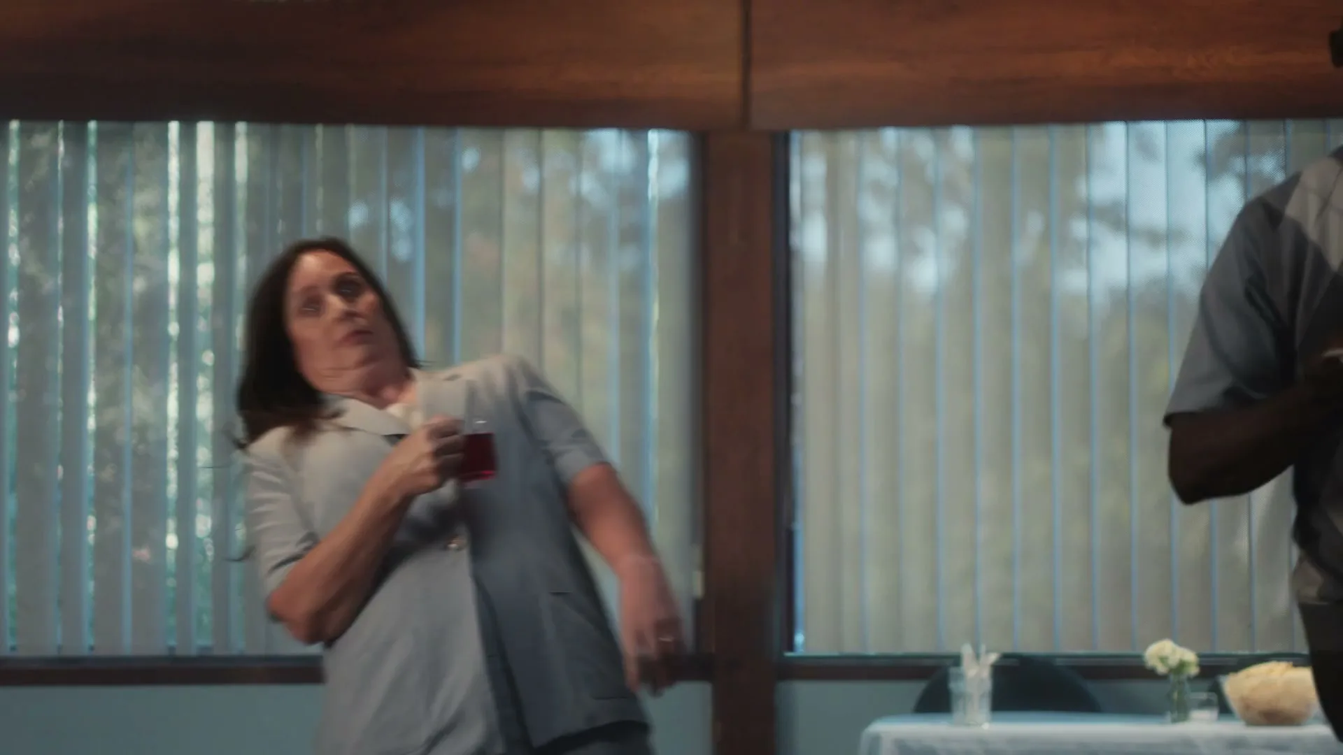 A woman in a light blue uniform holds a small glass of red liquid, looking confused and swaying as if affected by something, in a room with large windows.