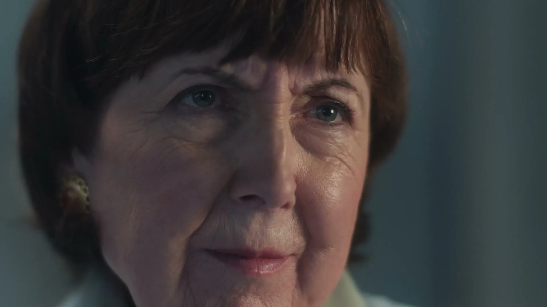 A close-up shot of an older woman with short brown hair, looking up and smiling slightly in an interior setting.