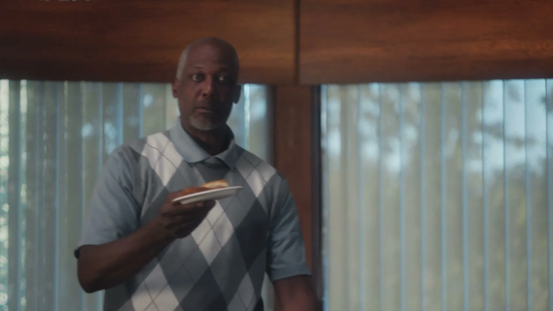 A man in his 50s, wearing a light blue argyle polo shirt, holds a small plate with food and looks around with a confused expression in a modern home.