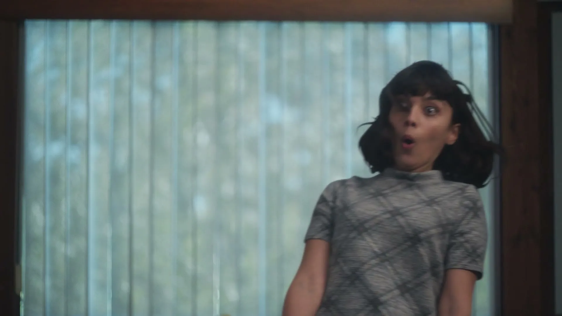 A young woman with dark hair and a patterned grey top looks surprised, with her mouth open, in an interior setting.
